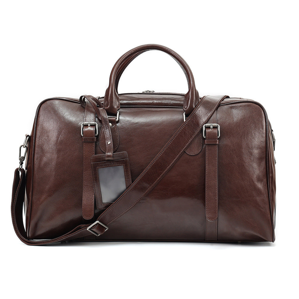 A dark brown leather duffel bag with dual top handles, a detachable shoulder strap, buckle details, and a small hanging luggage tag, shown against a plain white background.