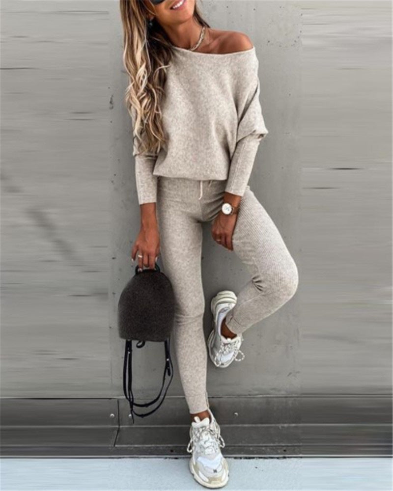 Woman wearing a gray off-shoulder top and matching pants, holding a black handbag against a gray wall.