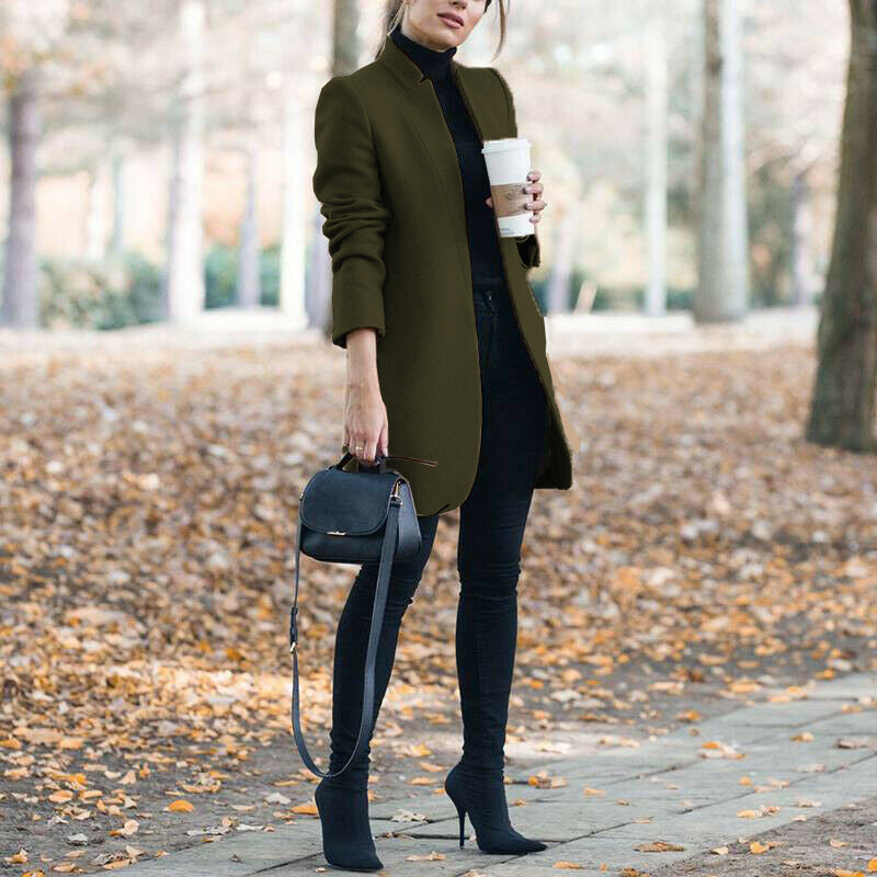 Woman in an olive green coat holding a coffee cup, standing on a leaf-covered path.