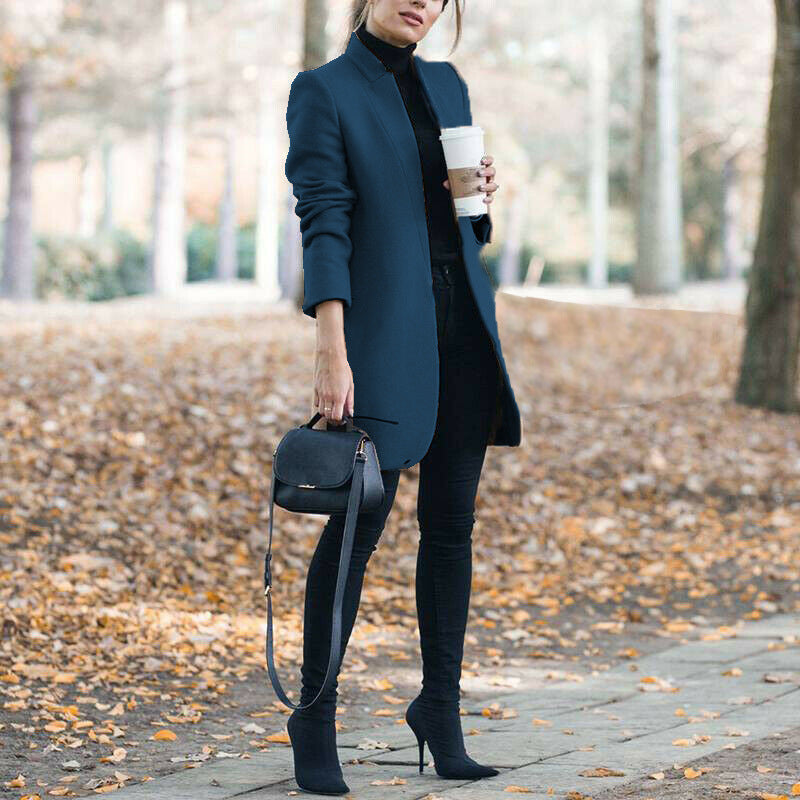 Woman in a blue coat holding a coffee cup, standing on a leaf-covered path.