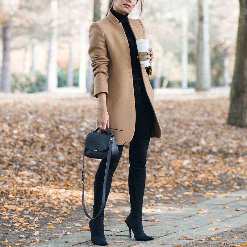 Woman in a tan coat holding a coffee cup, standing on a leaf-covered path.