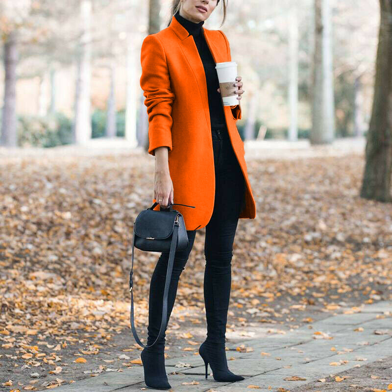 Woman in an orange coat holding a coffee cup in a park with fallen leaves on the ground.