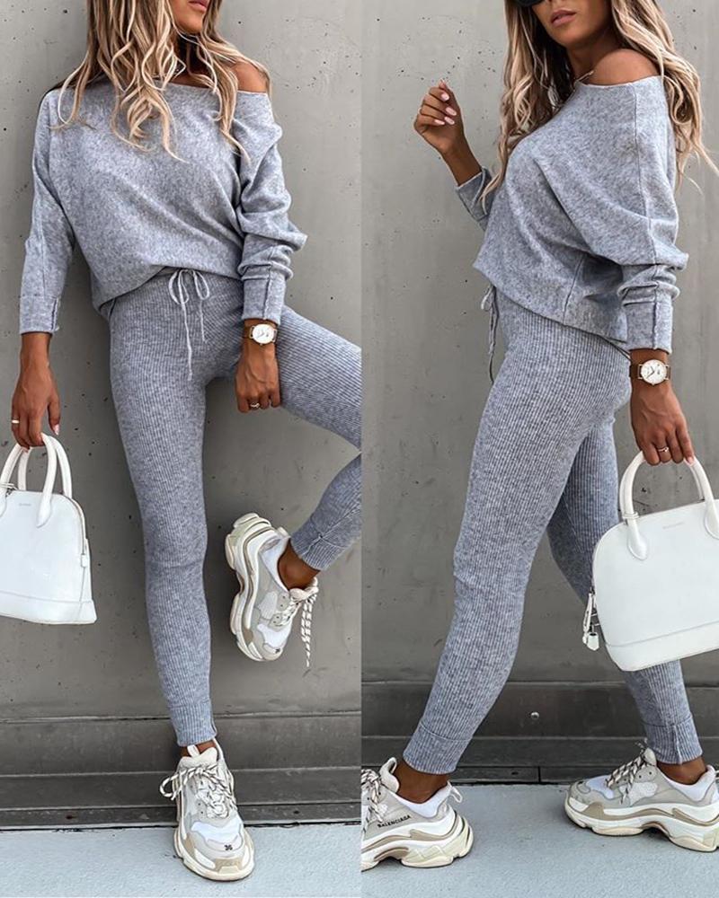 Woman wearing a gray tracksuit with white sneakers, holding a white handbag against a gray wall.