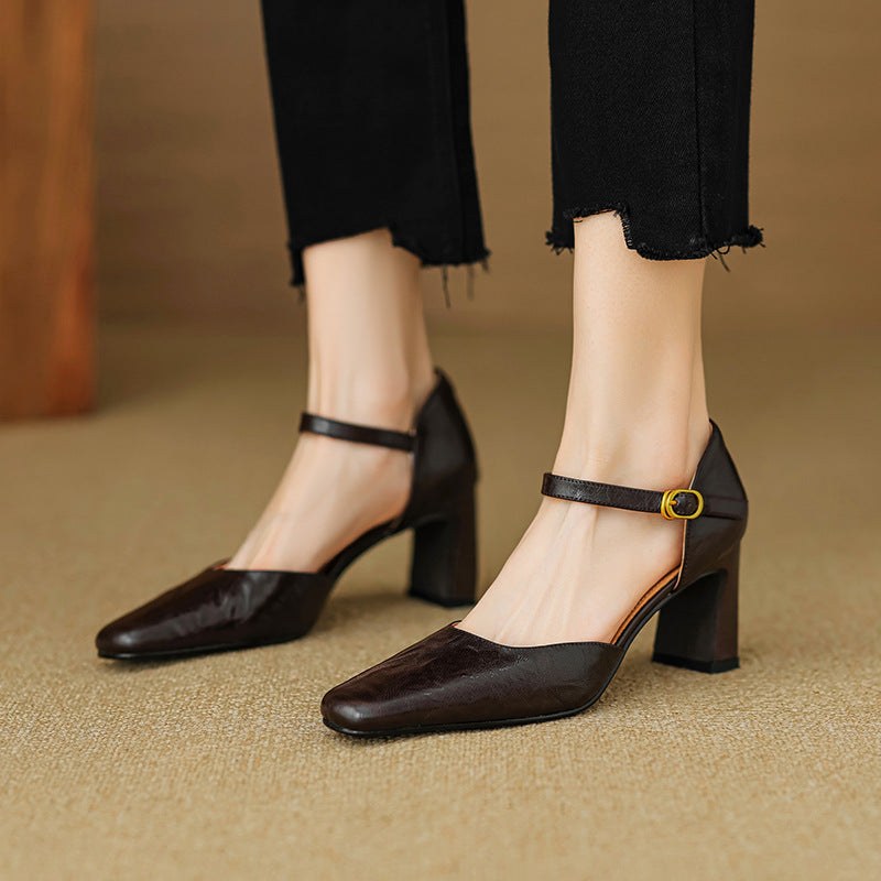 A pair of dark brown block-heel Mary Jane pumps with ankle straps and gold buckle details, shown on a model wearing black cropped jeans.