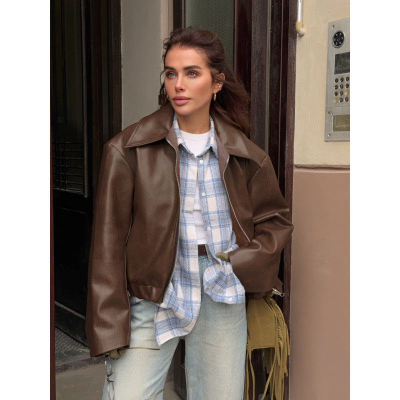 Woman wearing a brown leather jacket over a plaid shirt and jeans, standing in front of a door.