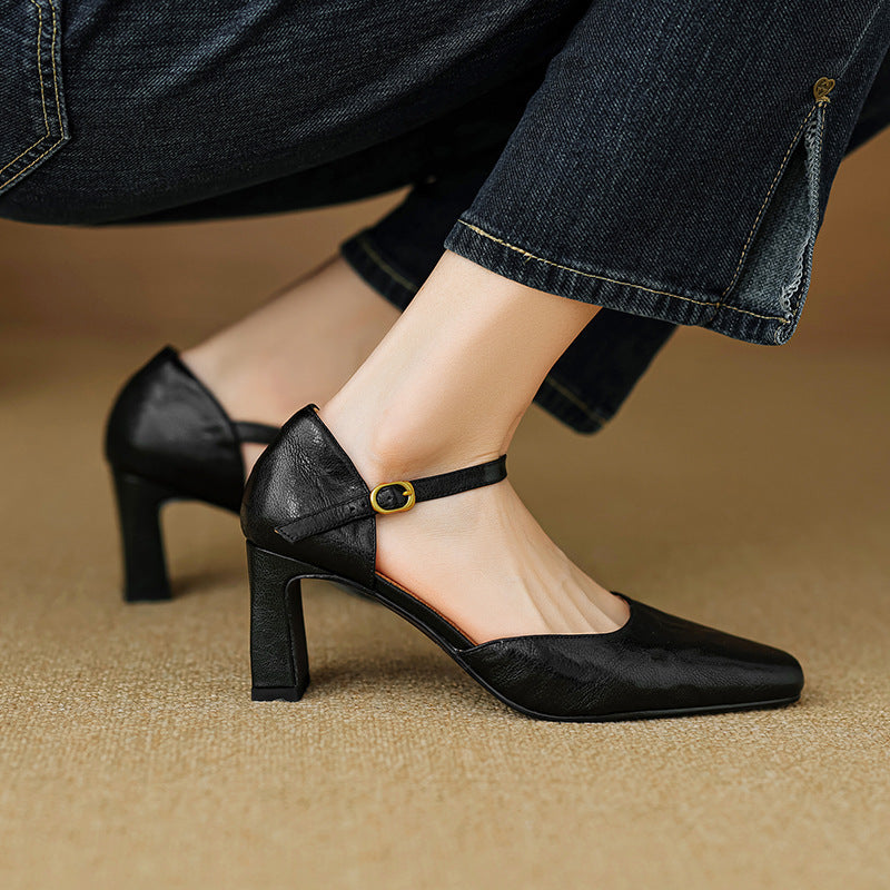 A close-up of a person wearing black leather block-heel pumps with ankle straps and gold-tone buckle details, paired with dark blue jeans, seated on a beige textured floor.