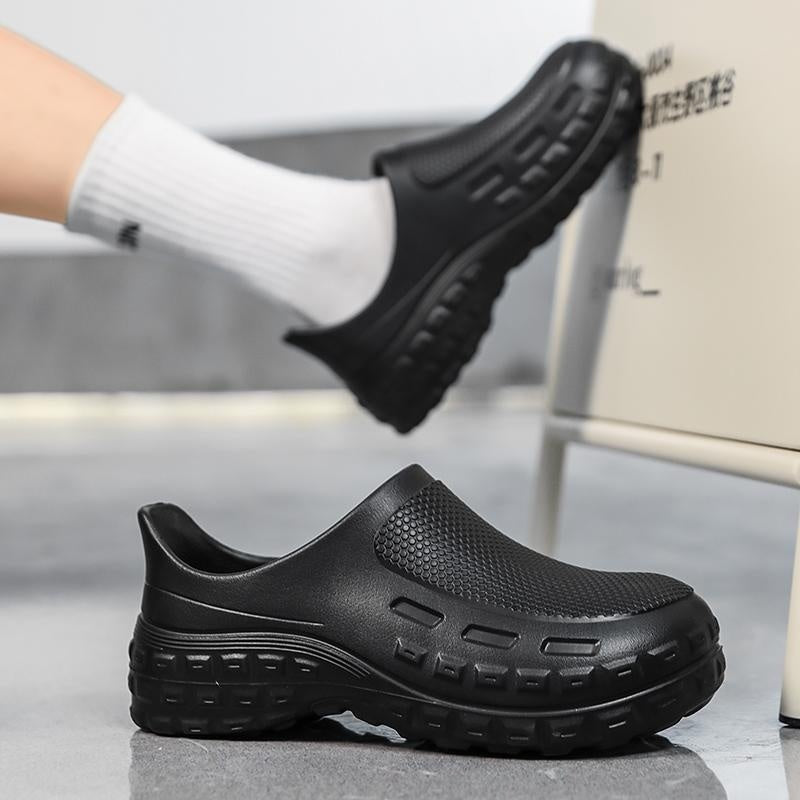 Black slip-on clog-style shoes with textured soles, shown with one shoe on a foot wearing a white sock and the other resting on the floor.