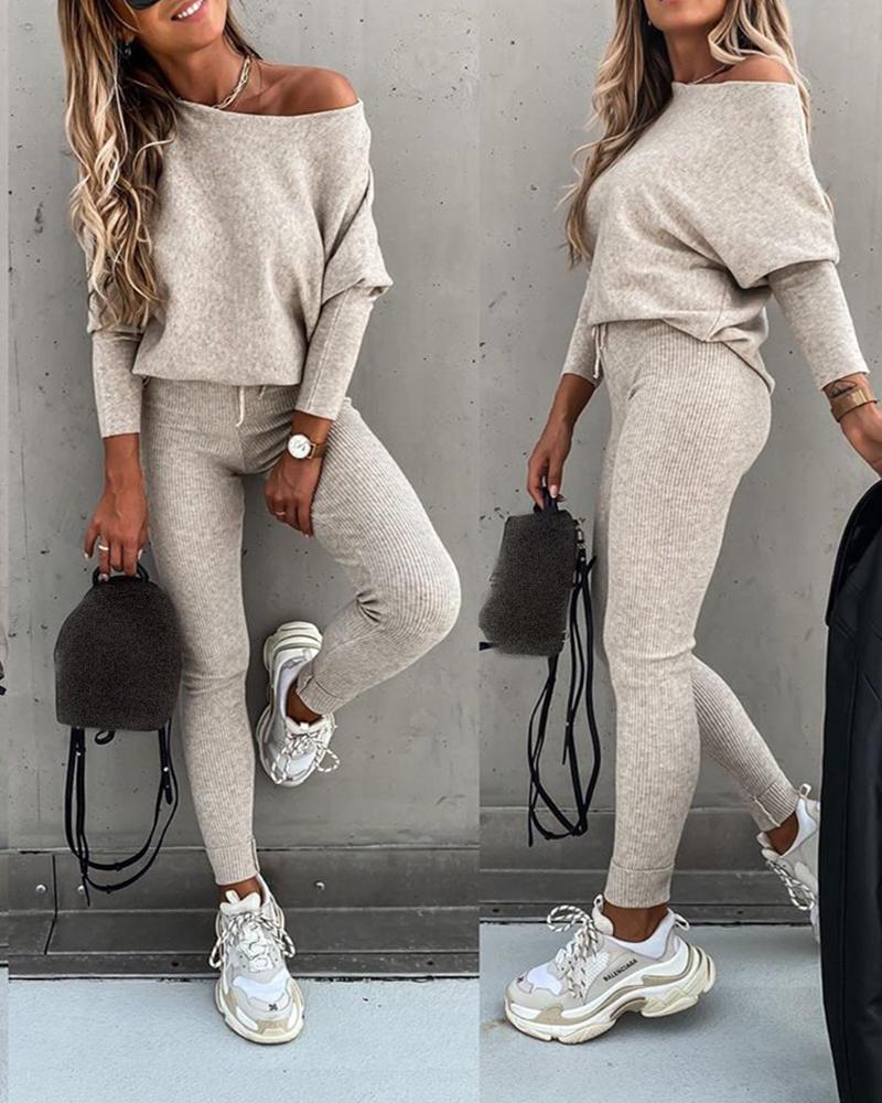 Woman wearing a gray off-shoulder top and pants with white sneakers, holding a black bag against a gray wall.