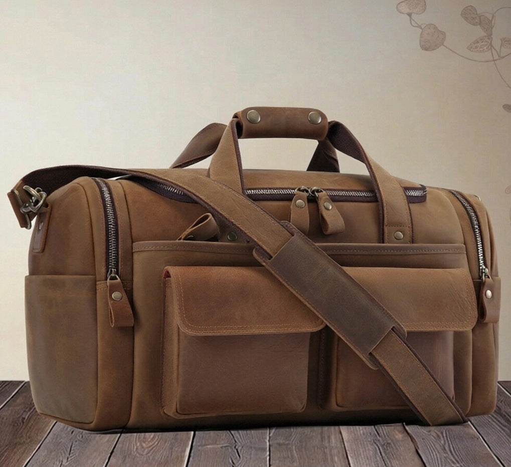A brown leather duffel bag with multiple zippered compartments, top handles, and a detachable shoulder strap, displayed on a wooden surface against a light beige wall.