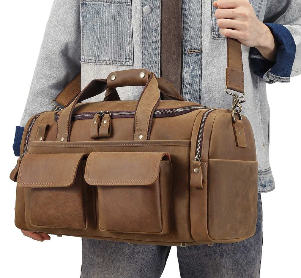 A brown leather duffel bag with multiple front pockets and top handles, shown being carried by a person wearing a denim jacket and jeans against a plain white background.