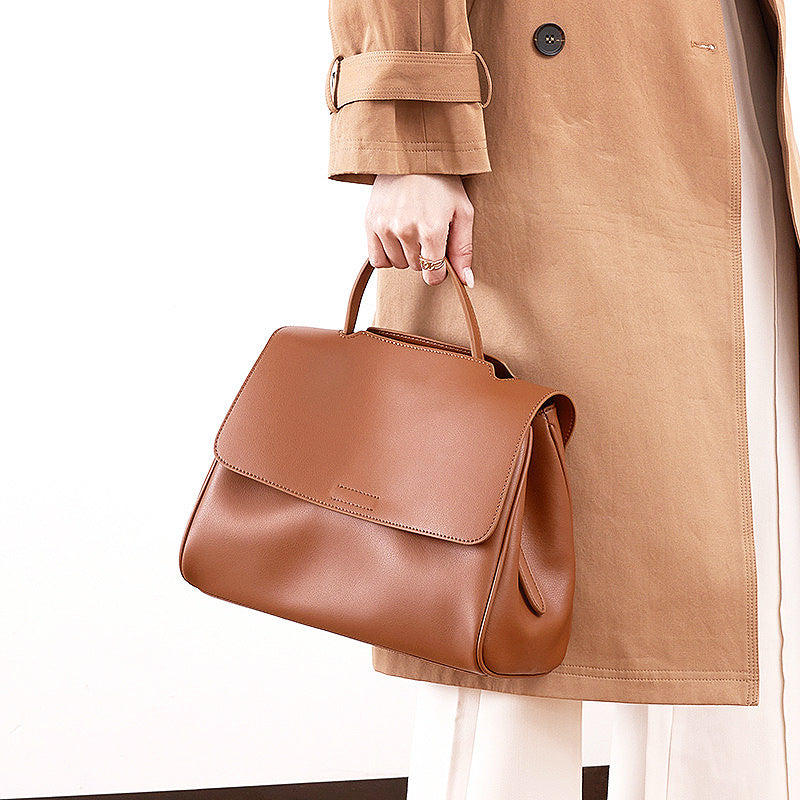 Brown leather handbag held by a person wearing a tan coat on a white background