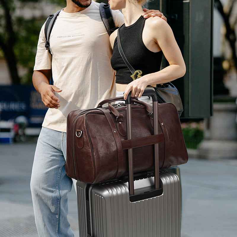 A man and a woman standing outdoors with a dark brown leather duffel bag resting on top of a silver rolling suitcase.