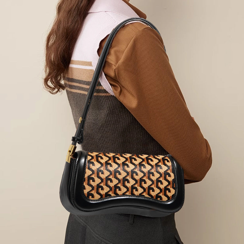A woman wearing a brown and beige striped sweater carries a black shoulder bag with a tan and black repeating monogram pattern on the front flap.