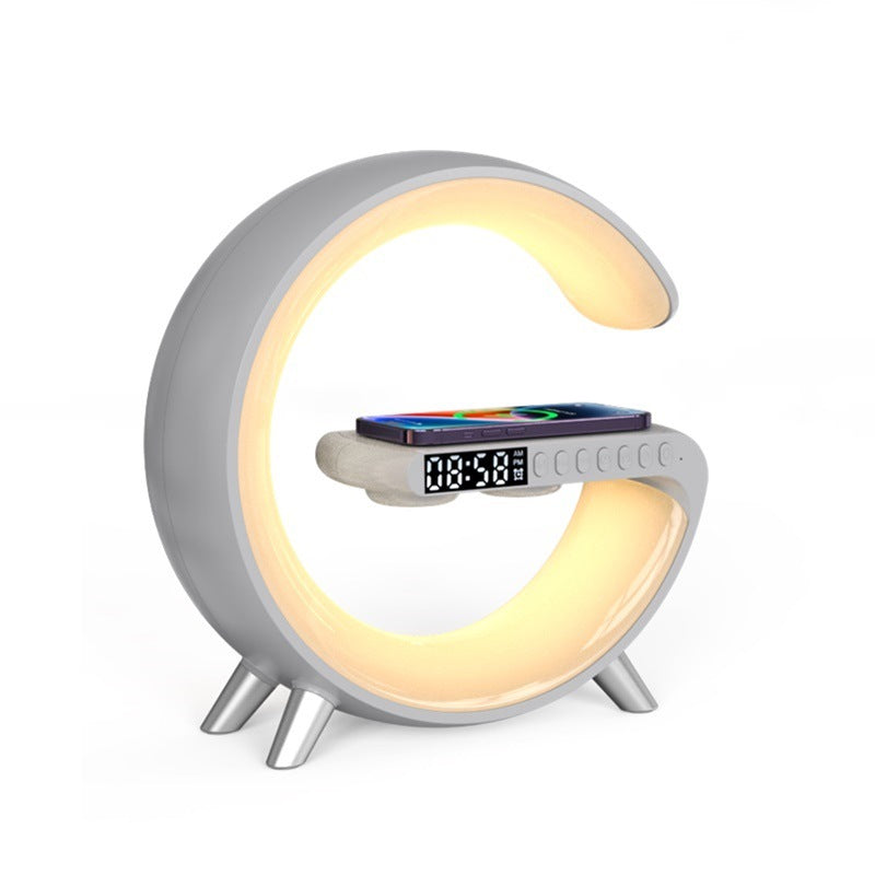 A modern C-shaped desk lamp with a built-in phone charger and digital alarm clock, featuring a warm yellow LED light glow and a silver-gray body on a white background.
