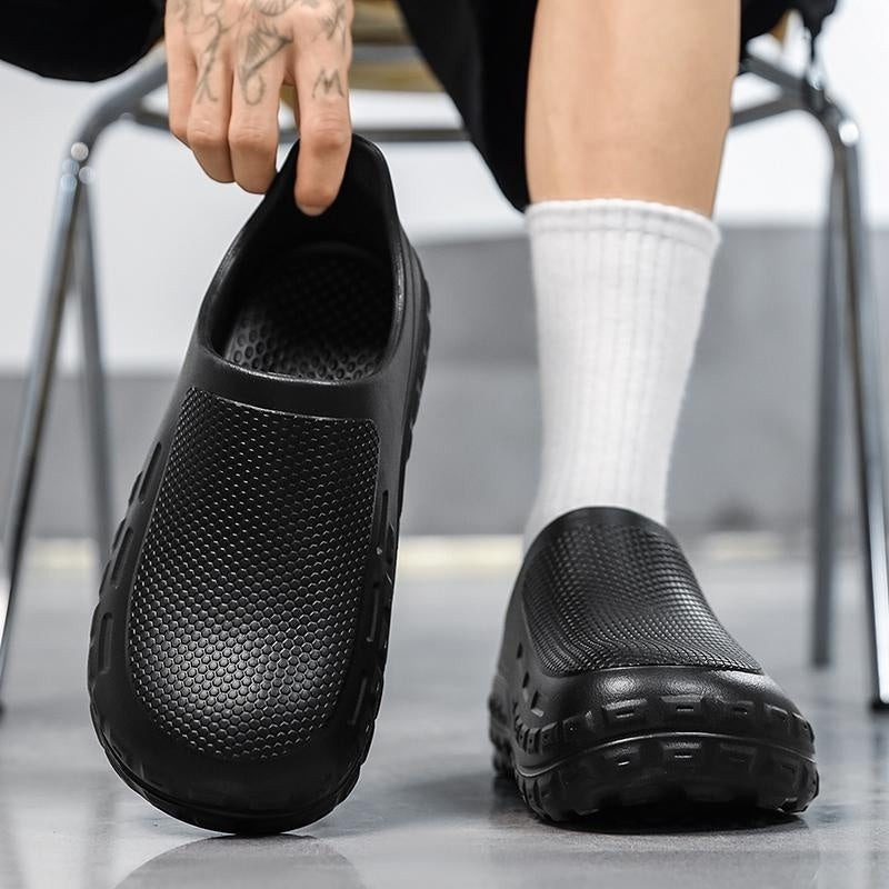 A person wearing black slip-on clog-style shoes with textured mesh and chunky lug soles, shown with one shoe being held by a tattooed hand and the other worn on a foot with a white sock.