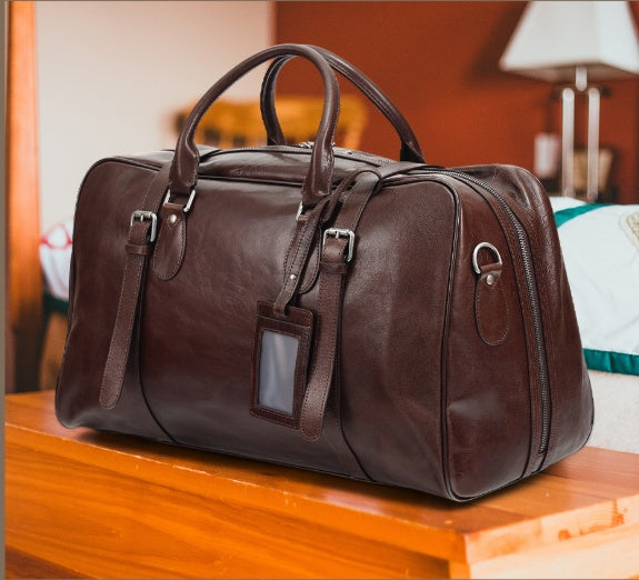 A dark brown leather duffel bag with dual top handles, buckle straps, and a luggage tag, resting on a wooden table in a warm-toned indoor setting.