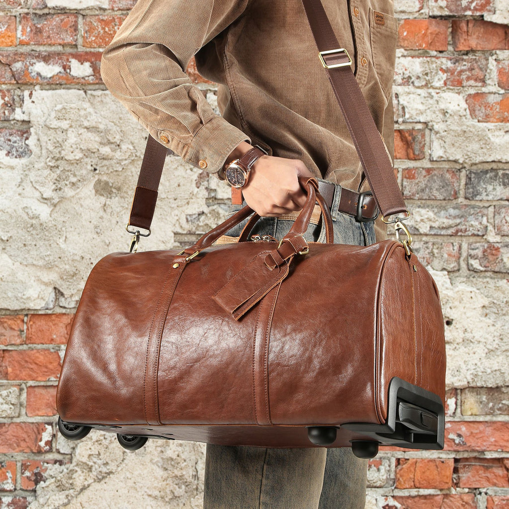 A brown leather duffel bag with a shoulder strap and wheels, held by a person wearing a brown corduroy shirt in front of a rustic brick wall.