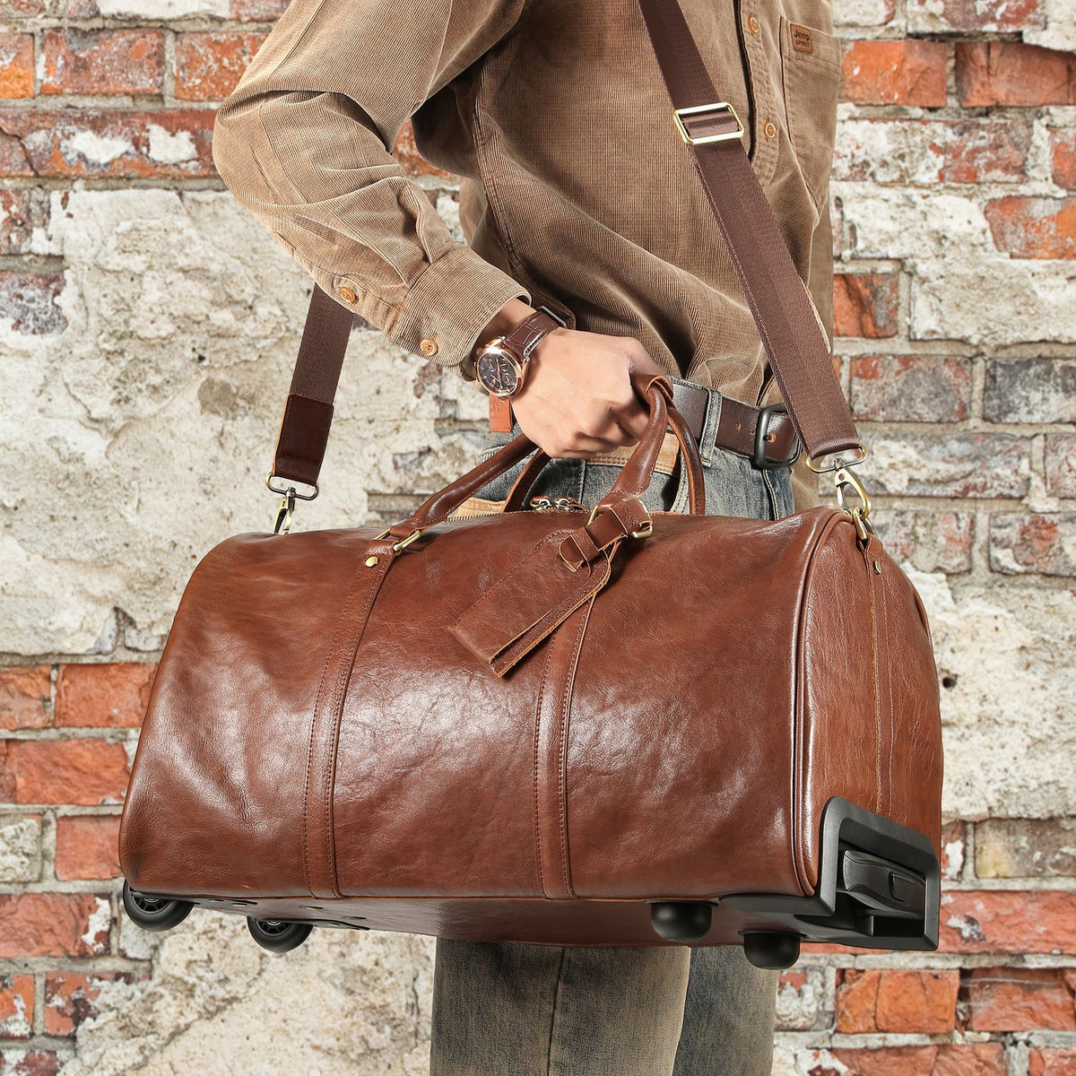 A brown leather duffel bag with a shoulder strap and wheels, held by a person wearing a brown corduroy shirt in front of a rustic brick wall.