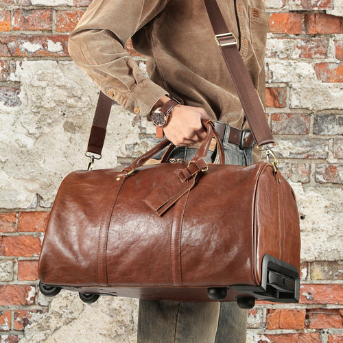 A brown leather duffel bag with a shoulder strap and wheels, held by a person wearing a brown corduroy shirt in front of a rustic brick wall.