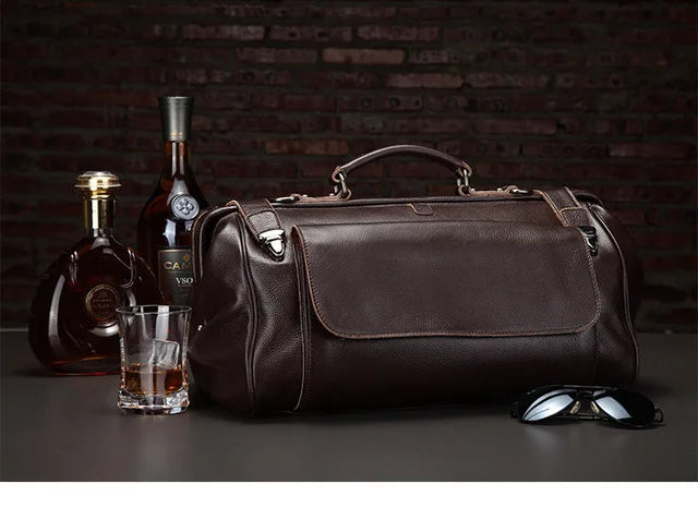 Brown leather duffel bag with a bottle of Cachél, a glass of whiskey, and sunglasses on a dark background.