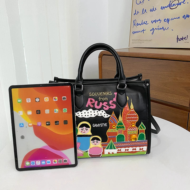 A black tote bag with double top handles and a colorful illustrated print reading 'SOUVENIRS from RUSSIA' and 'GAMASTE', placed on a white table next to an iPad. A real indoor setting with a dresser and wall is visible in the background, so this is Lifestyle rather than Product/Packaging.