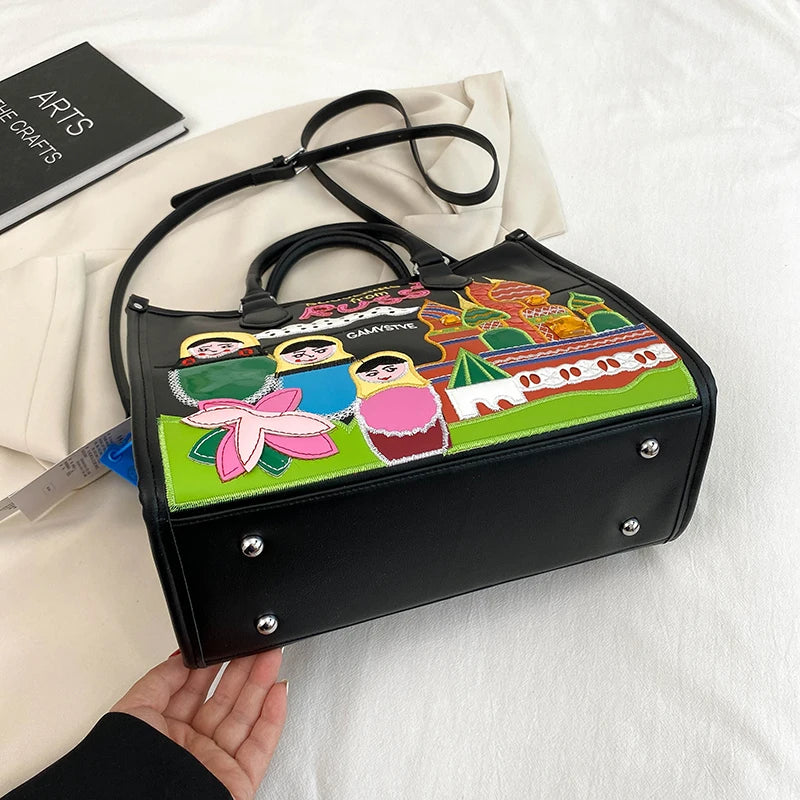 A black handbag with a colorful embroidered patch featuring traditional Russian nesting dolls and a pink lotus flower, resting on a white fabric surface next to a cream-colored dust bag.
