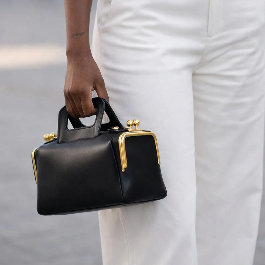 Black handbag with gold accents held by a person wearing white pants.