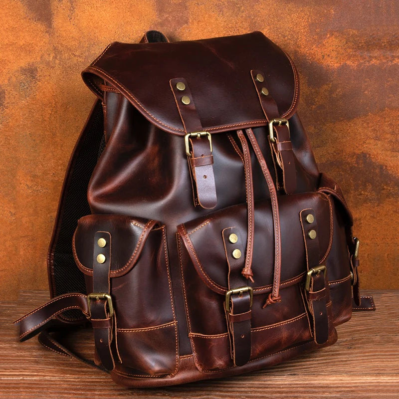 A dark brown leather backpack with multiple front pockets, brass-tone buckles, and drawstring closure, photographed on a wooden surface against a rusted metal background.