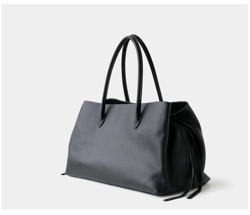 A black pebbled leather tote bag with dual top handles and a drawstring detail at the base, photographed on a clean light gray background.