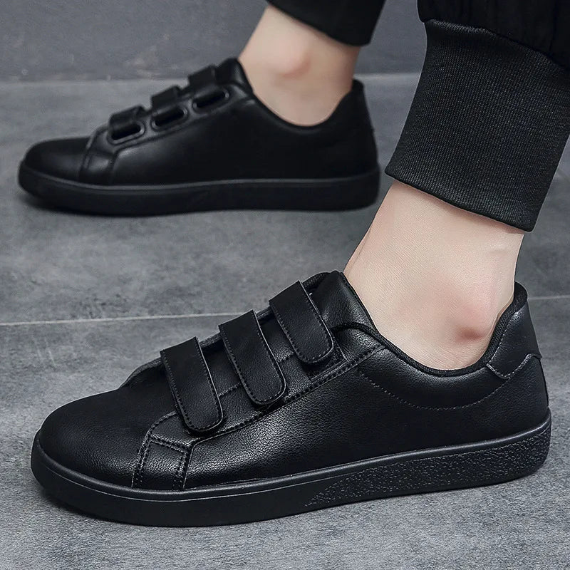 Black sneakers with velcro straps worn by a person on a gray floor.