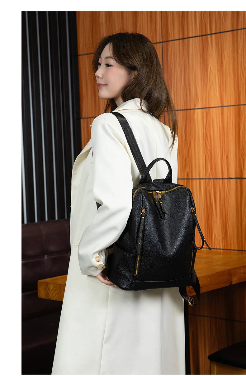 A woman wearing a long cream coat carries a black pebbled leather backpack with gold-tone hardware and a top handle, standing indoors against a wood-paneled wall.