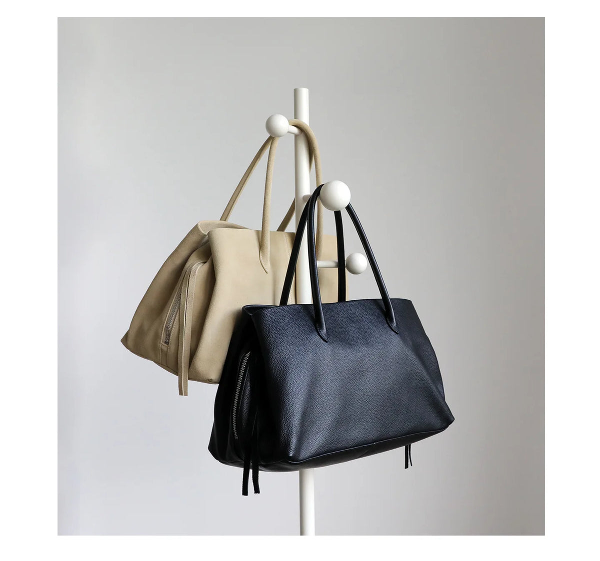 Two handbags, one beige and one black, hanging on a white stand against a light gray background.