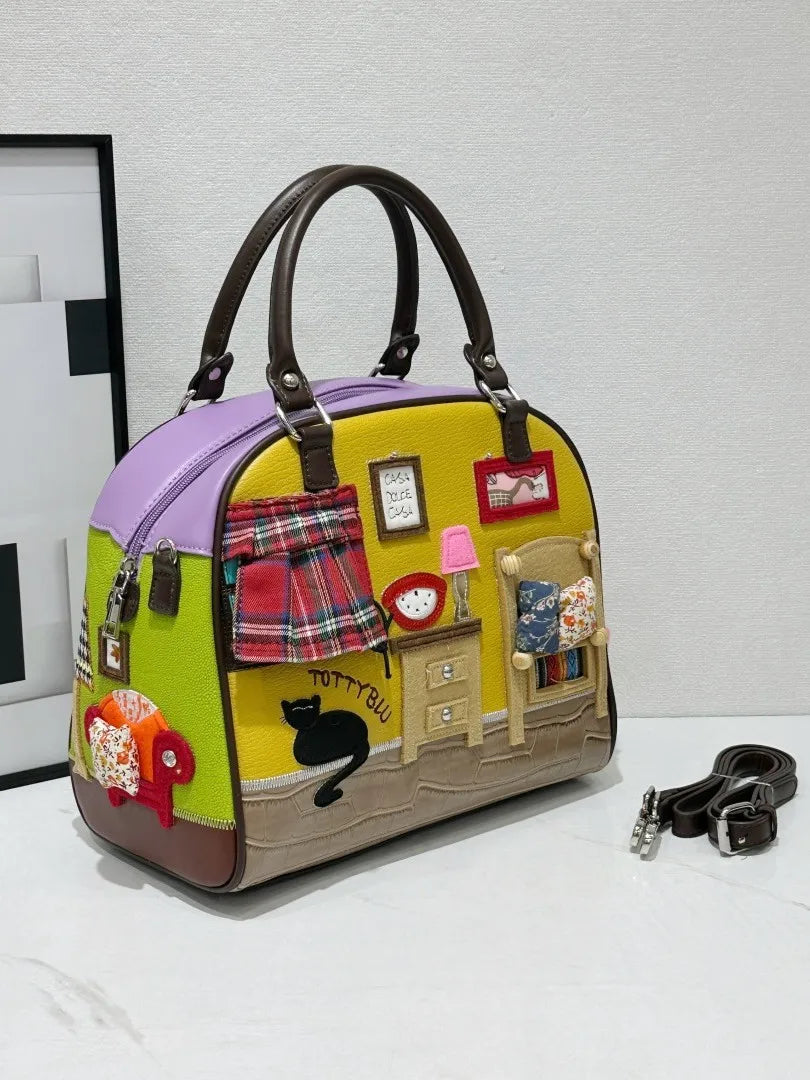 A whimsical dome-shaped handbag with a colorful patchwork design inspired by a cozy room scene. The bag features a yellow front panel with a miniature bed, dresser, and lamp, a purple top section, and brown leather handles and trim.