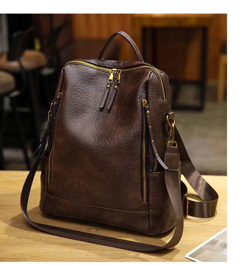 A dark brown faux-leather backpack with gold-tone zippers and hardware, displayed on a wooden table in a softly blurred indoor setting.