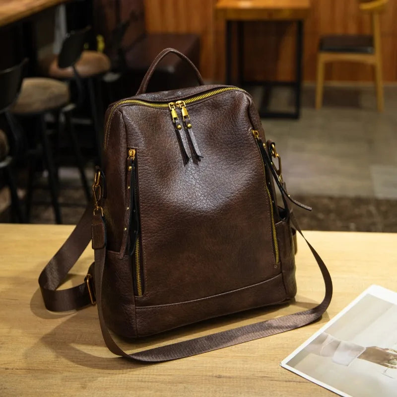 A dark brown textured leather backpack with gold-tone zippers and hardware, displayed on a light wooden table in a cafe setting.