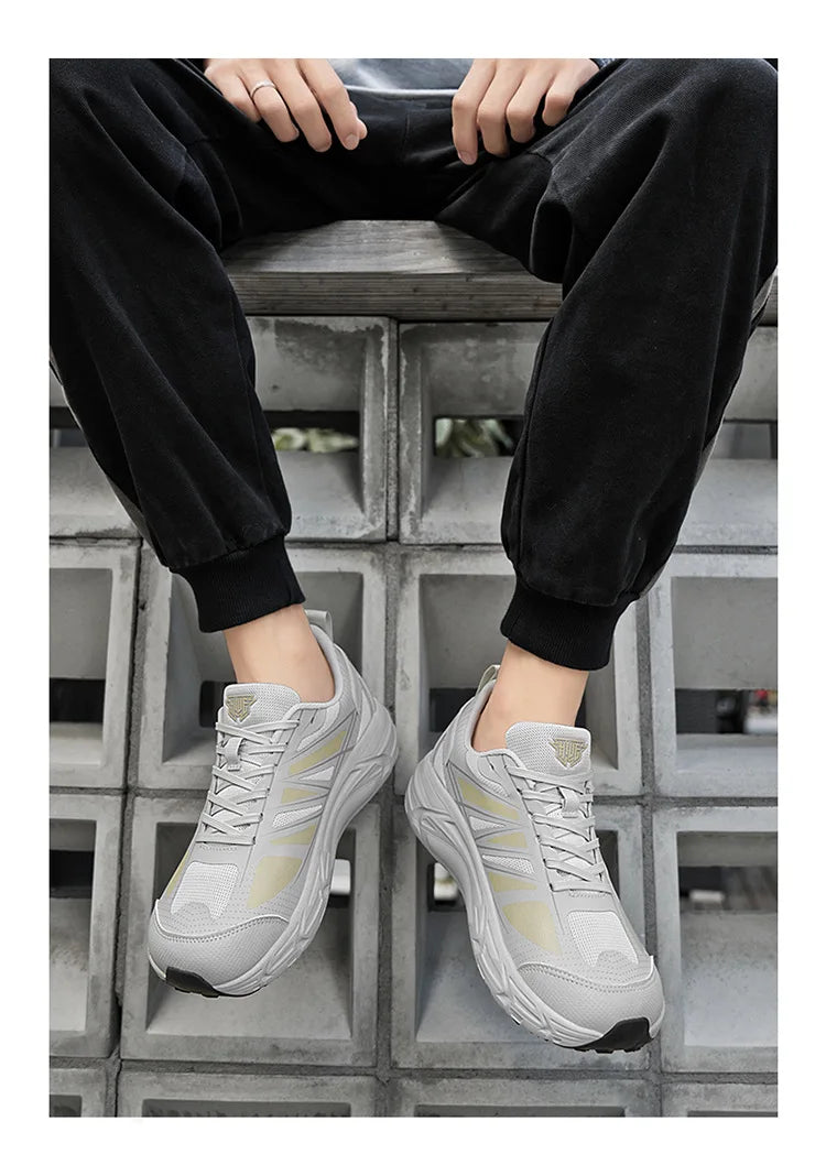 A person sitting on a concrete block wearing a pair of chunky low-top sneakers in light gray and white with subtle gold accents, paired with black jogger pants.