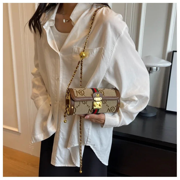 A person wearing an oversized white shirt carries a small Gucci shoulder bag with a beige monogram canvas body, brown leather trim, a red and green web stripe, and a gold chain strap.