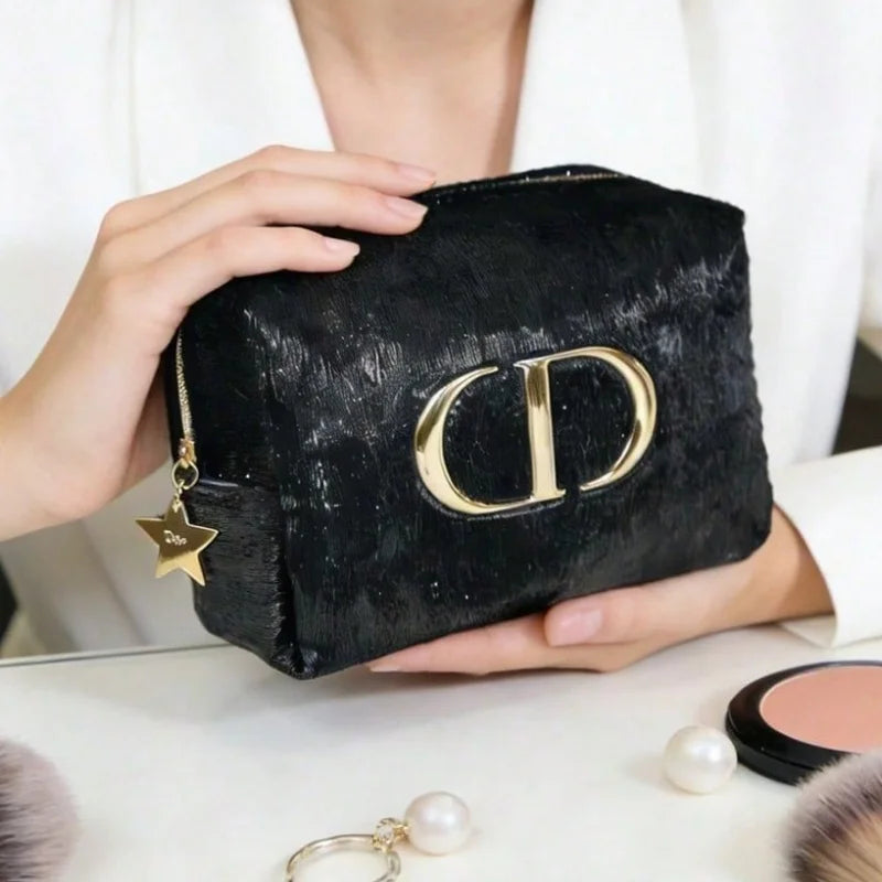 A person holds a black textured cosmetic pouch with a large gold-tone CD logo and a star charm, shown in a close-up lifestyle shot with makeup items on a table.