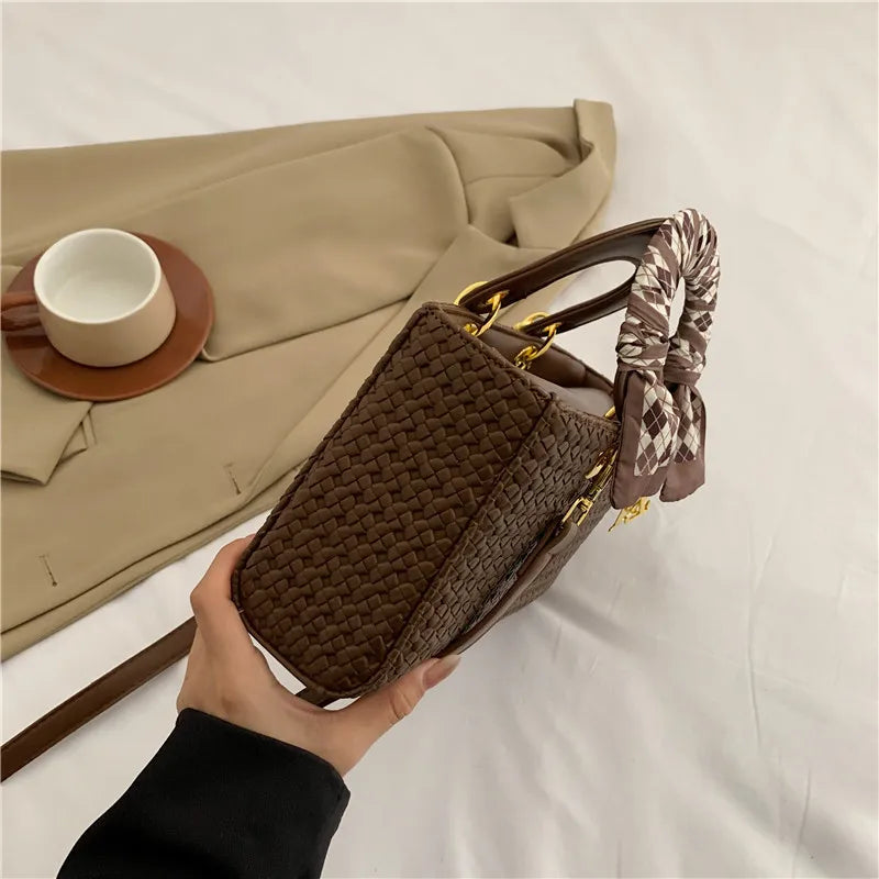 Brown woven handbag held by a hand with a beige surface and cup in the background