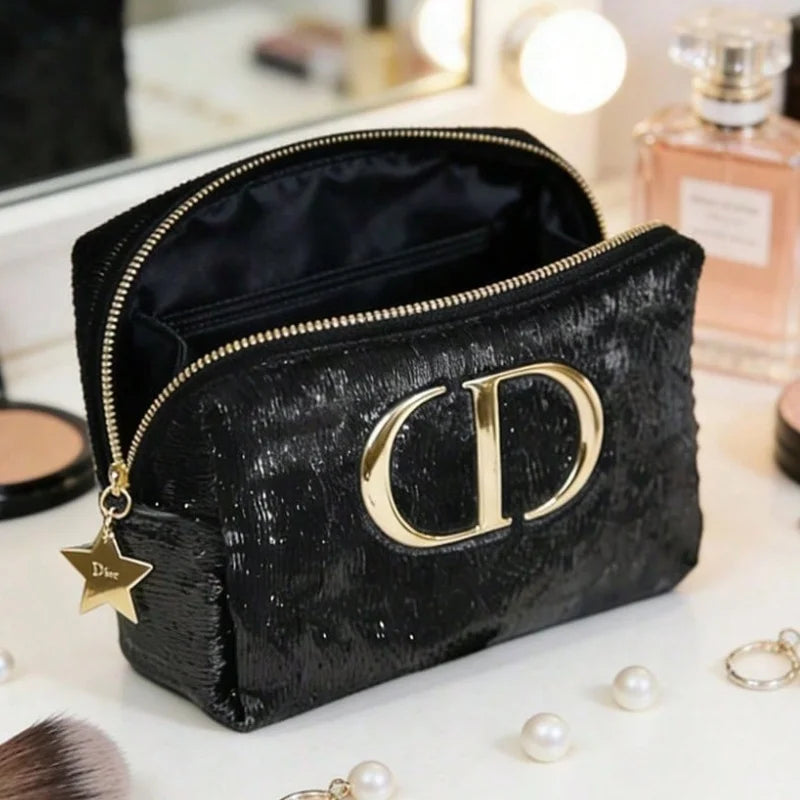A black textured cosmetic pouch with a large gold-tone 'CD' logo on the front, gold zipper, and a dangling star charm reading 'Cde', shown on a vanity surface with perfume and makeup items in the background.