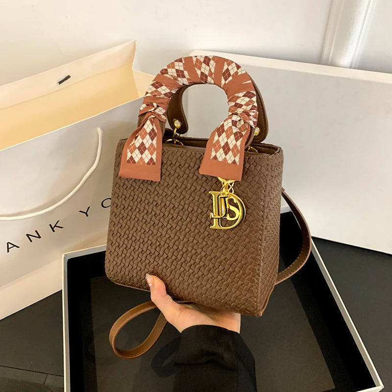 Brown woven handbag with a patterned strap held by a hand, with a brand logo on a white background.