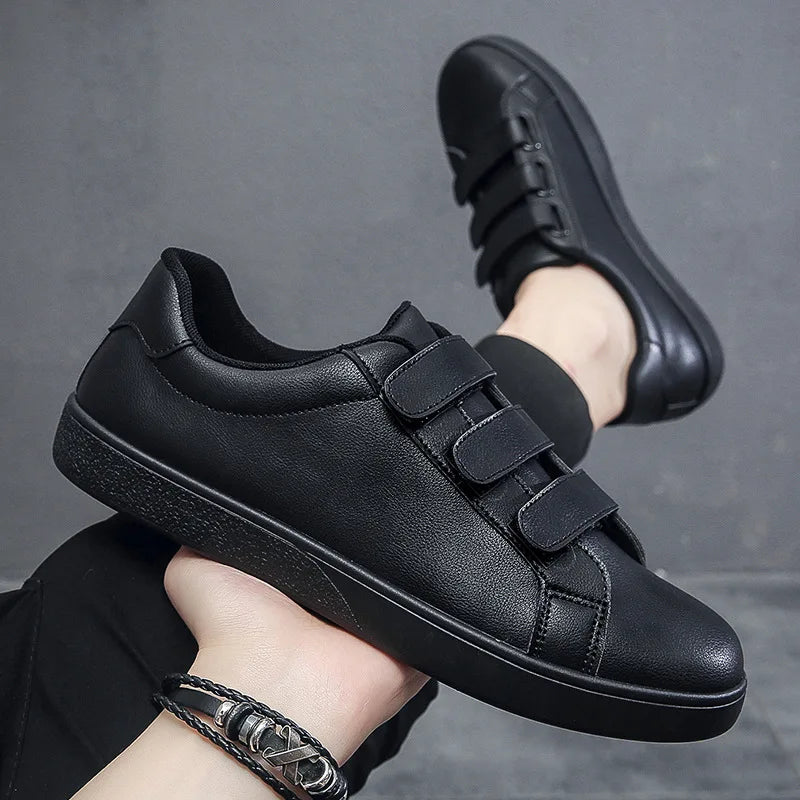 Black sneakers with velcro straps held by a person against a gray background