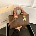 Brown textured handbag with a decorative handle held by a person, with a white surface and beige bag in the background.