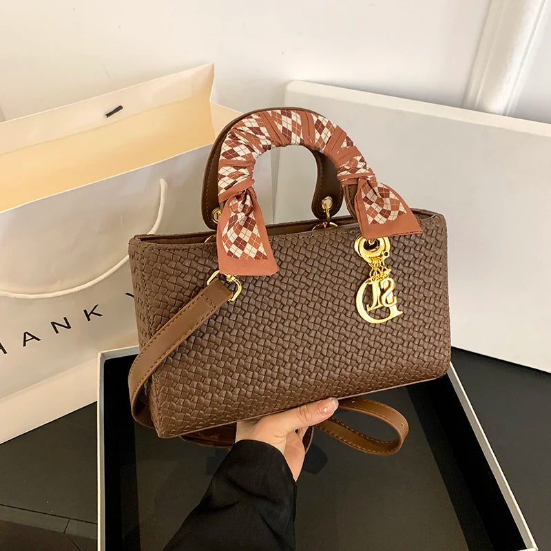 Brown textured handbag with a decorative handle held by a person, with a white surface and beige bag in the background.