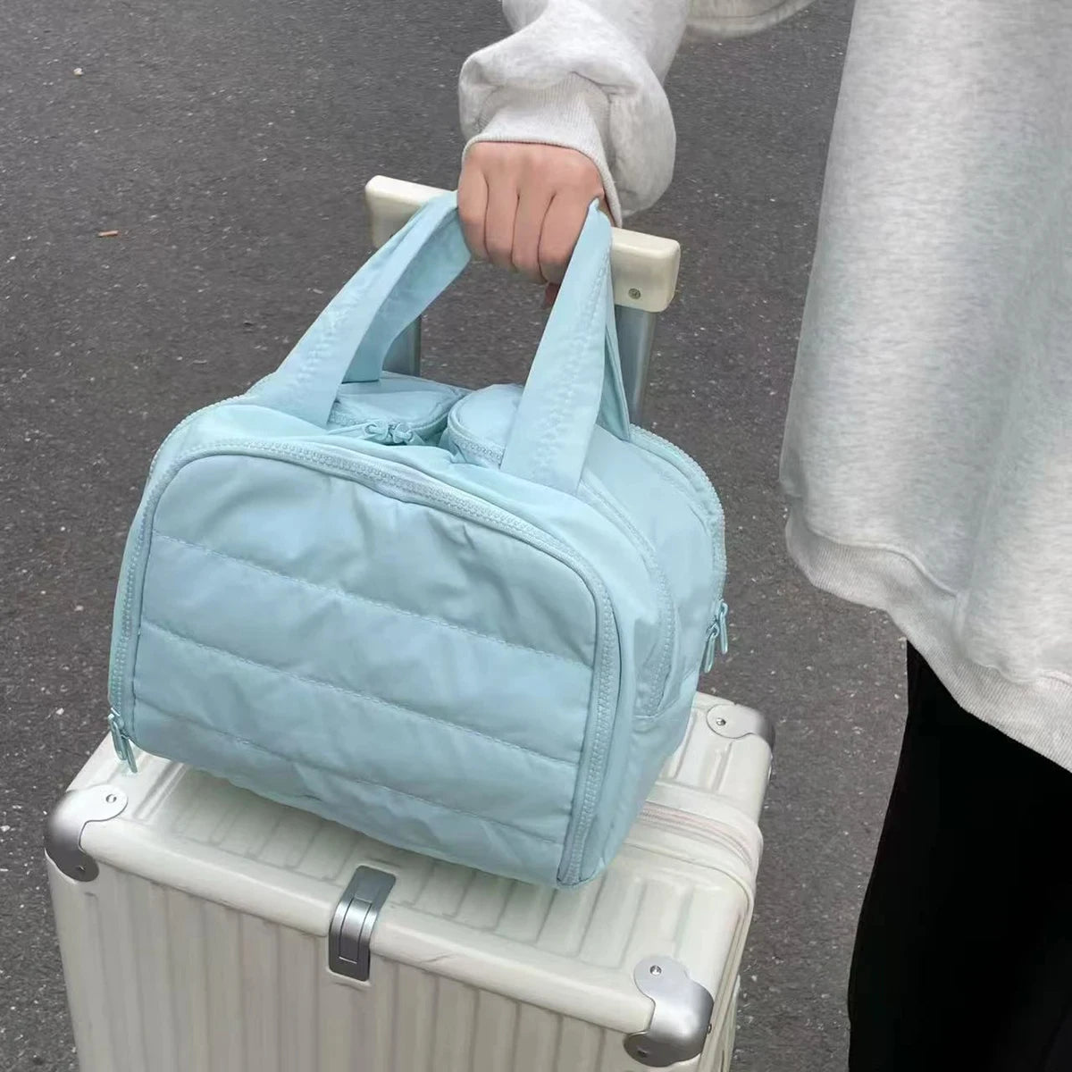 Light blue quilted bag being held above a white suitcase on a gray pavement.