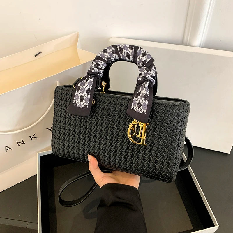 Black woven handbag with a patterned handle held by a hand, with a white box in the background.