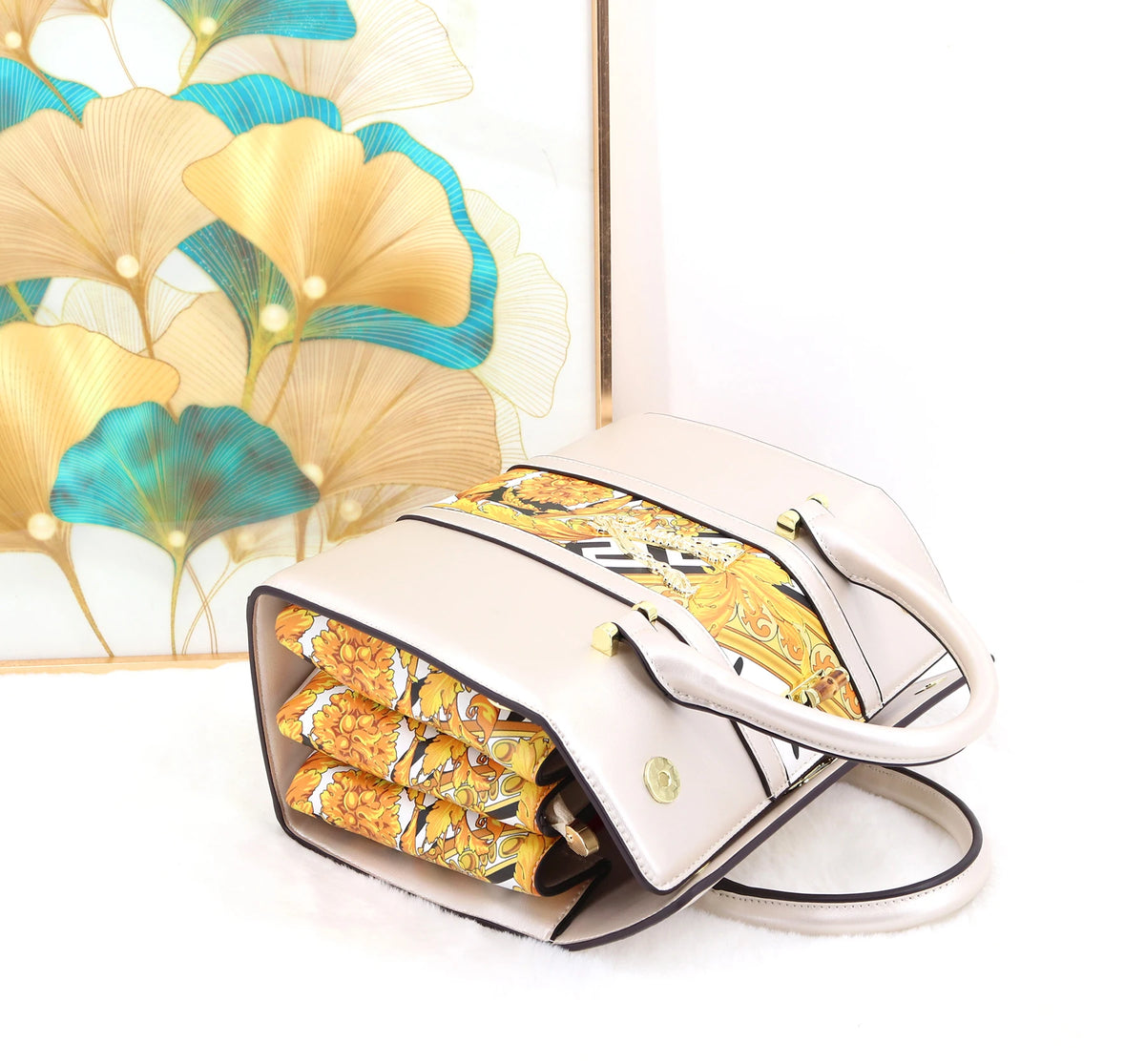 A structured beige handbag with a gold and black patterned scarf draped across the top and tucked inside, set against a white background with a decorative gold and teal leaf print frame.