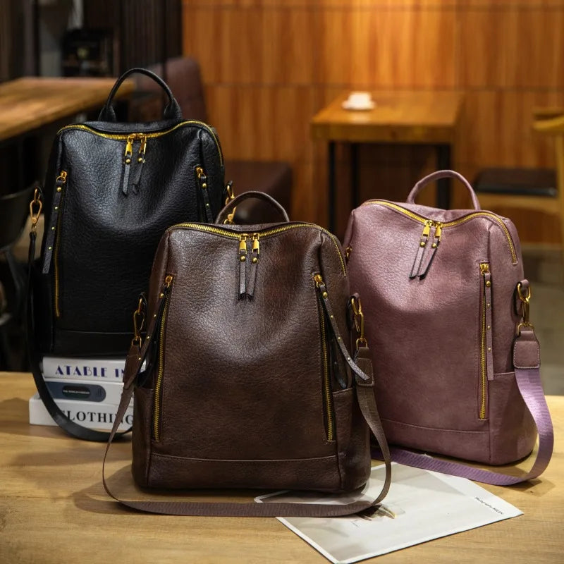 Three pebbled leather backpacks in black, dark brown, and dusty mauve are arranged on a wooden table in a warm, wood-paneled interior setting. Each backpack features gold-tone zippers, dual top handles, and a detachable shoulder strap.