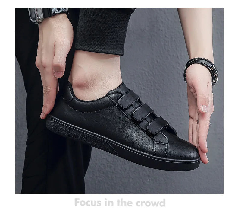 Black casual shoe with velcro straps worn by a person on a gray background