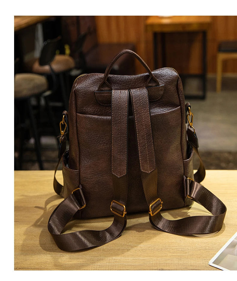 A dark brown textured leather backpack with adjustable shoulder straps and gold-tone hardware, displayed on a light wooden table in a cafe setting.